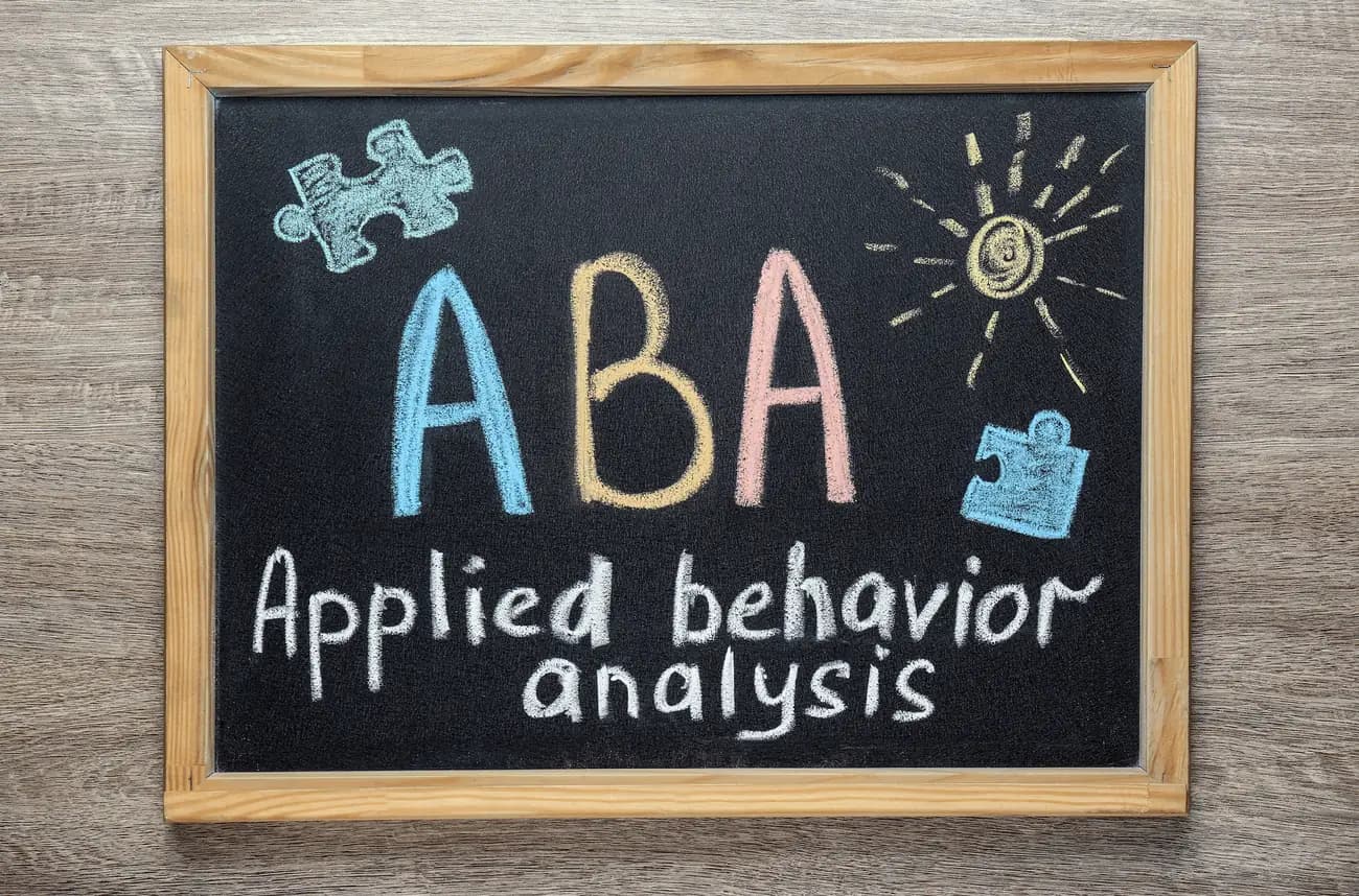 A chalkboard with the letters ABA and the words Applied Behavior Analysis written in coloured chalk, surrounded by puzzle-piece drawings