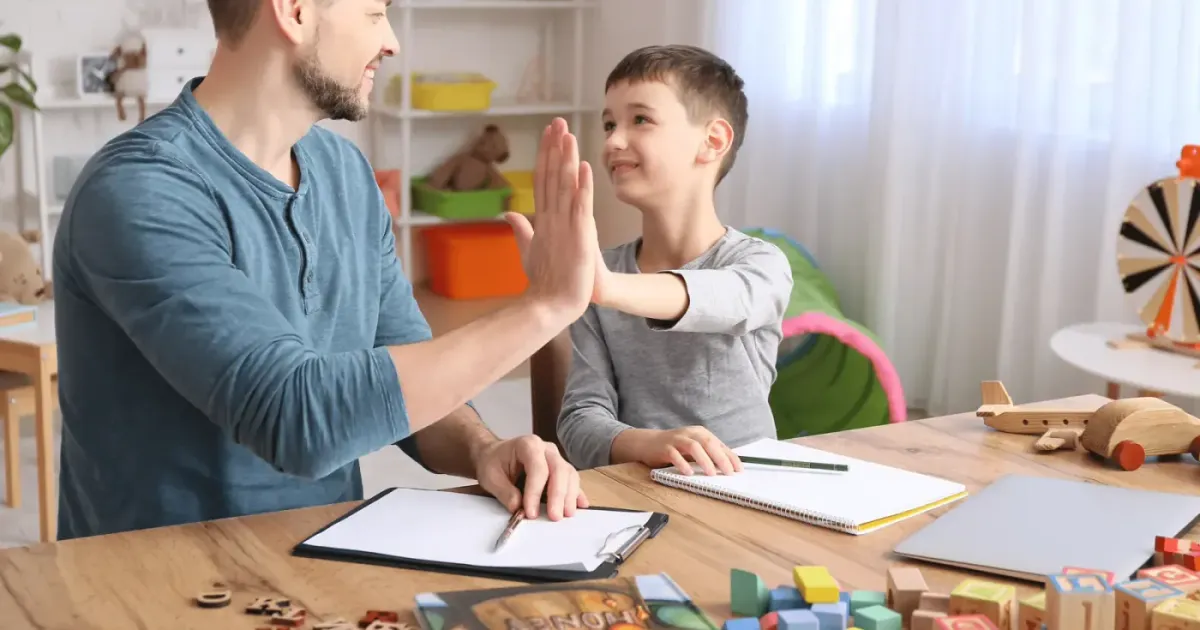 A therapist and a child high-fiving across a table during a session, with session notes and a clipboard in front of them