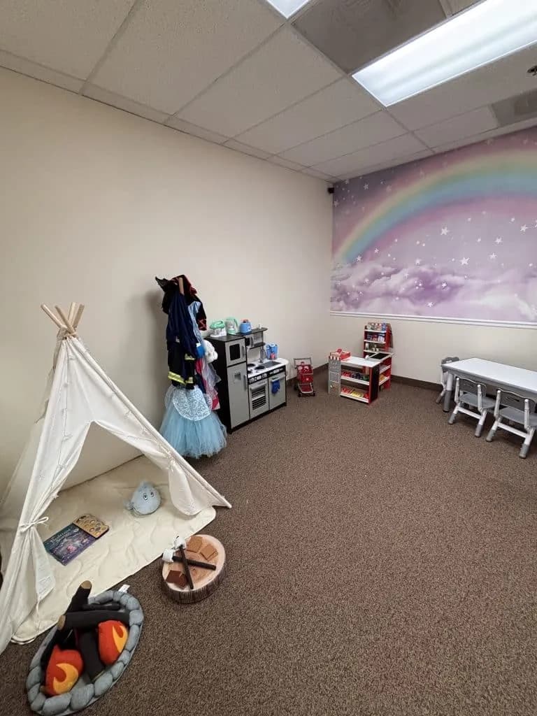 An imaginative-play room with a canvas teepee, play kitchen, dress-up rack, rainbow wall mural and a child-height table with chairs