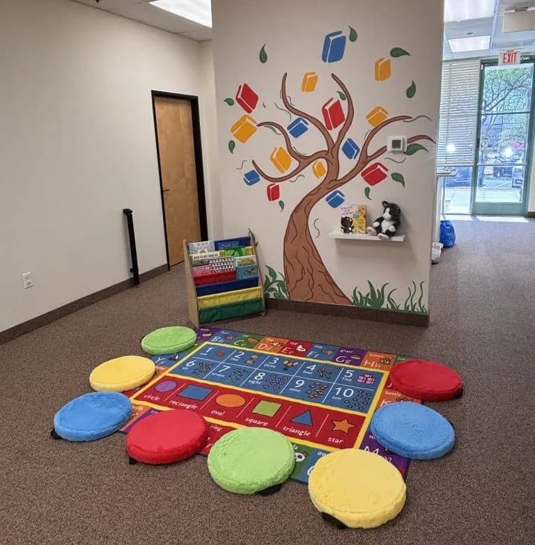A reading corner with a painted tree mural full of colourful books, a low book rack, and an alphabet and numbers rug ringed with round floor cushions