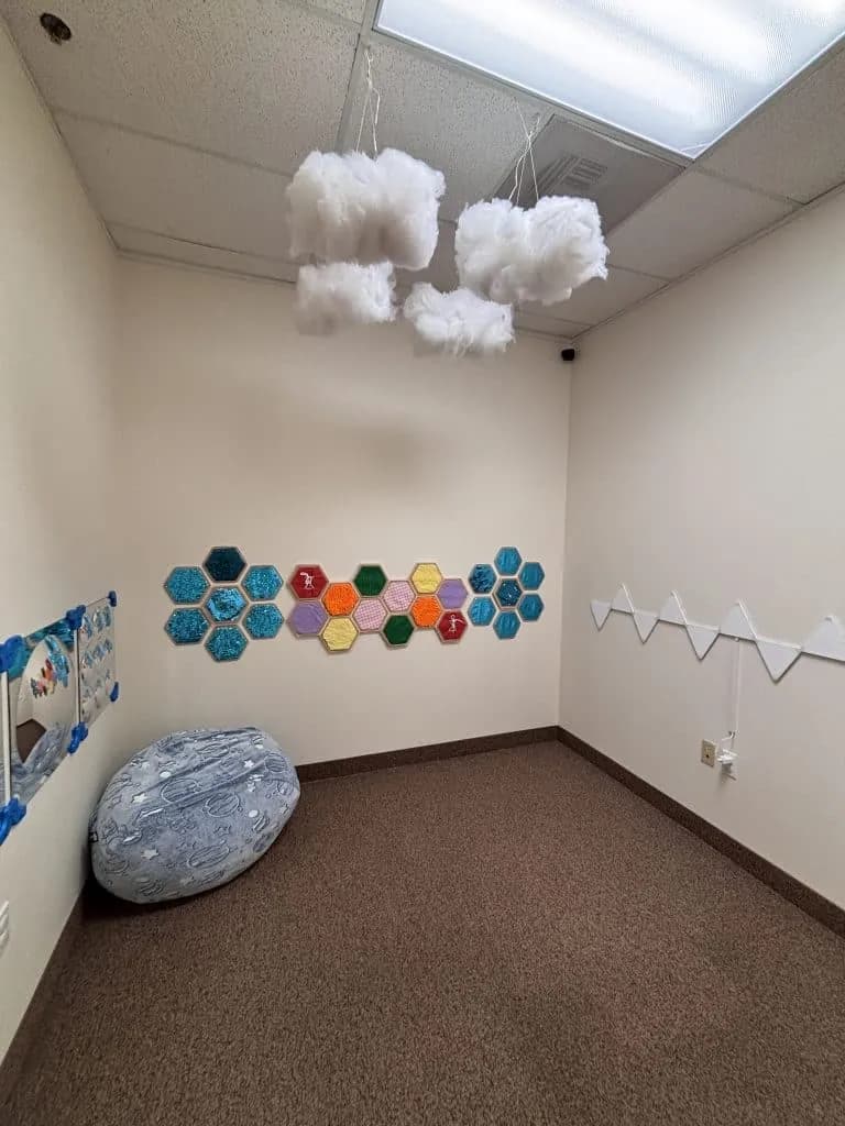 A calm sensory room with textured hexagon wall panels, cotton cloud mobiles hanging from the ceiling, a large bean bag and a floor mirror