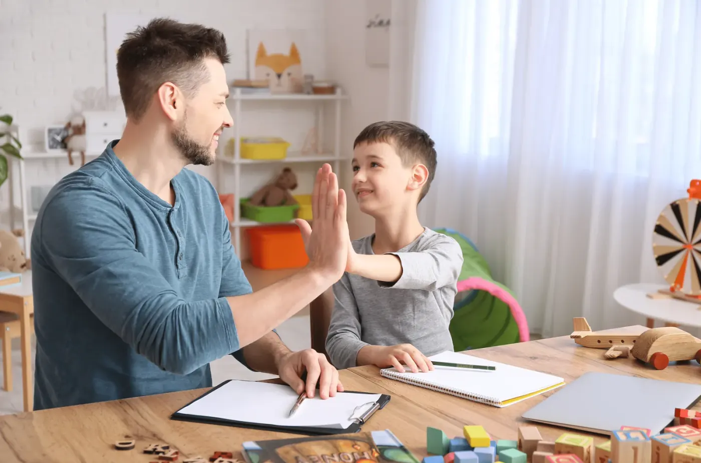 A therapist and a child high-fiving across a table during a session, with session notes and a clipboard in front of them